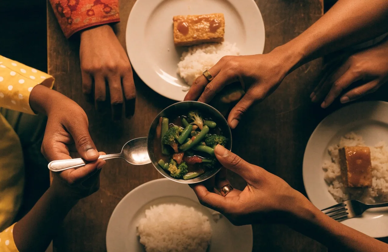 A family at a simple dinner table passing a bowl of vegetables alongside rice and tempeh